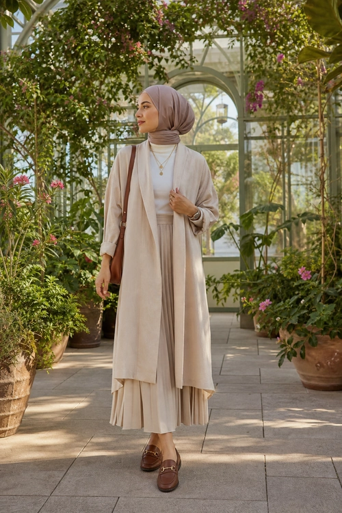 A woman wearing a taupe hijab, cream long cardigan, light top, pleated skirt, brown shoulder bag, and brown loafers in a greenhouse.