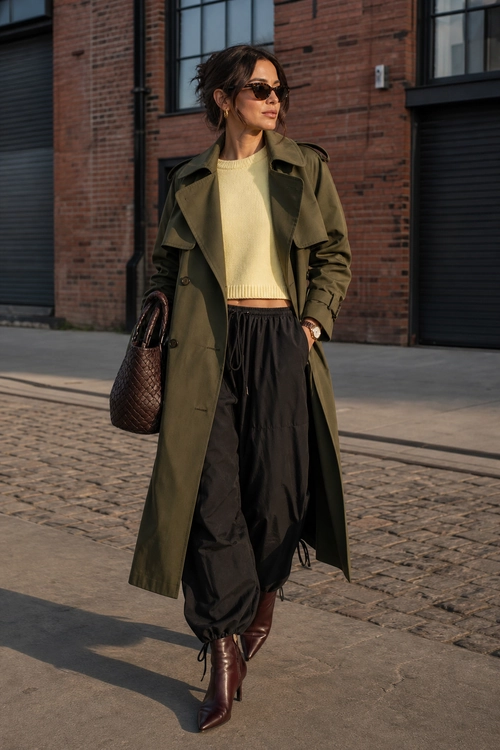 Street style outfit featuring an olive trench coat, cream cropped sweater, black drawstring trousers, burgundy pointed boots, sunglasses, and a woven brown tote bag.