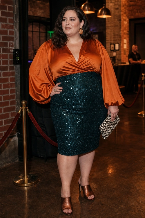 Orange Blouse and Teal Sequin Skirt A woman wears an orange satin wrap blouse with voluminous sleeves, a teal sequin skirt, brown heeled sandals, gold earrings, and a woven clutch.