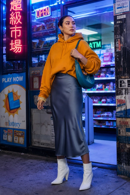 An orange hoodie styled with a dark gray satin midi skirt, teal shoulder bag, and white ankle boots.