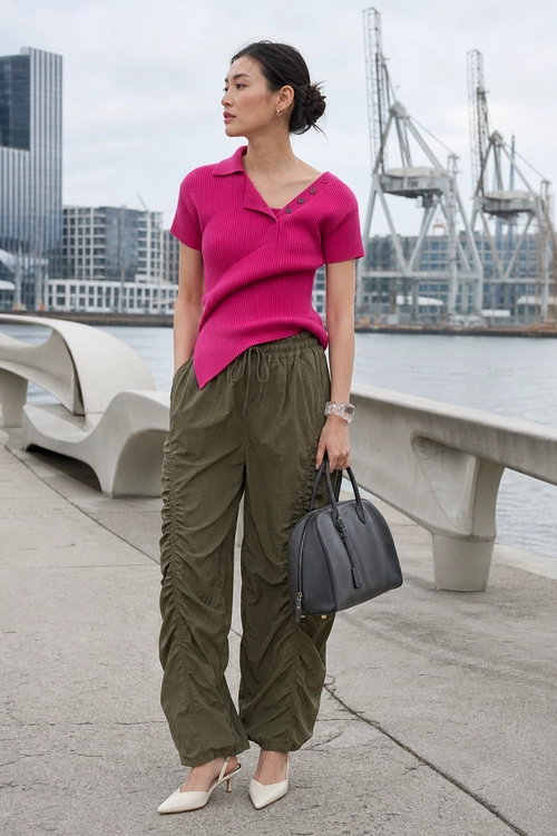 A woman wears a bright pink short-sleeve knit top with olive ruched trousers, cream pointed heels, and a dark handbag.