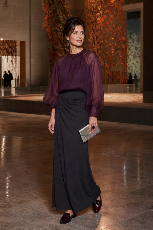 A woman wears a sheer plum blouse with voluminous sleeves, a draped black floor-length skirt, burgundy loafers, and carries a silver clutch.