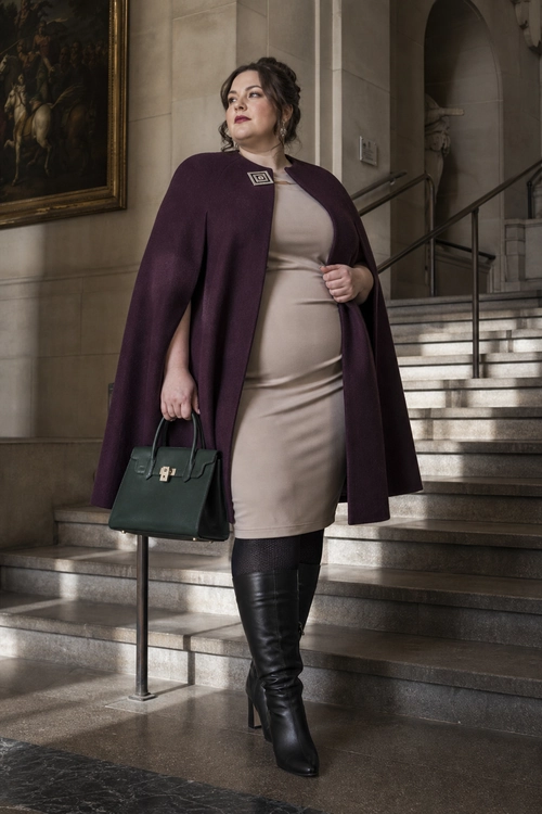 Plum Cape and Beige Dress Outfit A woman wears a plum cape over a beige fitted dress with black over-the-knee boots and carries a dark green handbag.