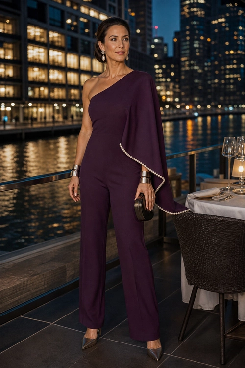 Woman wearing a plum one-shoulder jumpsuit with a draped cape sleeve, pearl trim, metallic pumps, cuff bracelets, drop earrings, and a black clutch.