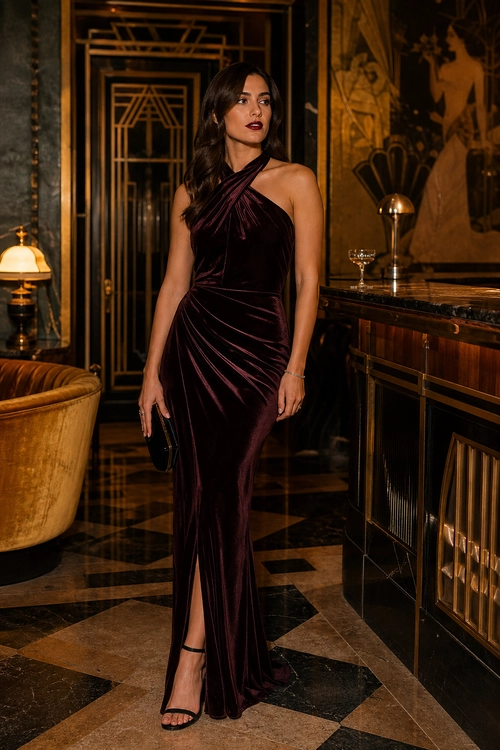 Plum Velvet Evening Gown Woman wearing a draped plum velvet halter gown with a thigh-high slit, black strappy heels, and a small black clutch.