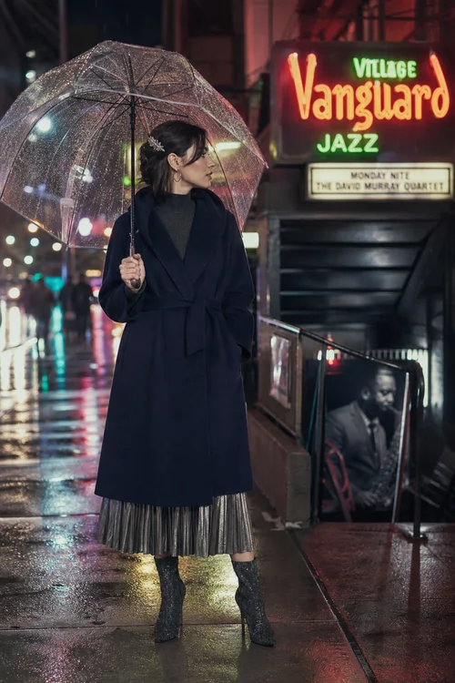 A woman holds a clear umbrella while wearing a dark belted coat, gray turtleneck, metallic pleated skirt, and black ankle boots on a rainy night.