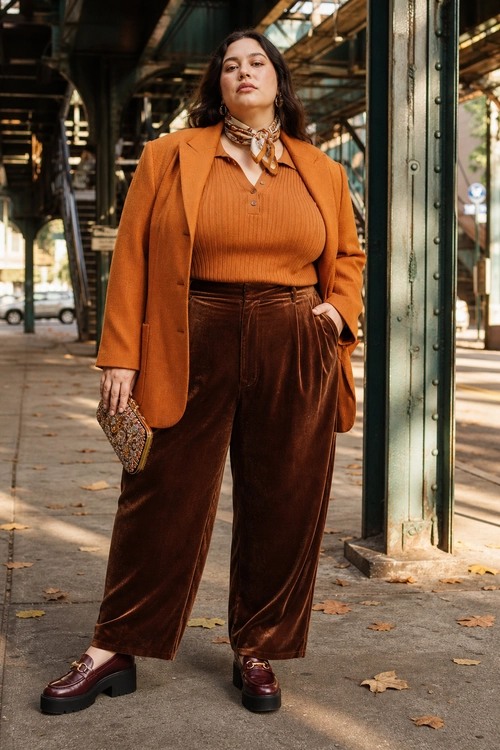 A woman wears a rust blazer over a ribbed knit top with brown velvet wide-leg trousers, a patterned neck scarf, and burgundy platform loafers.