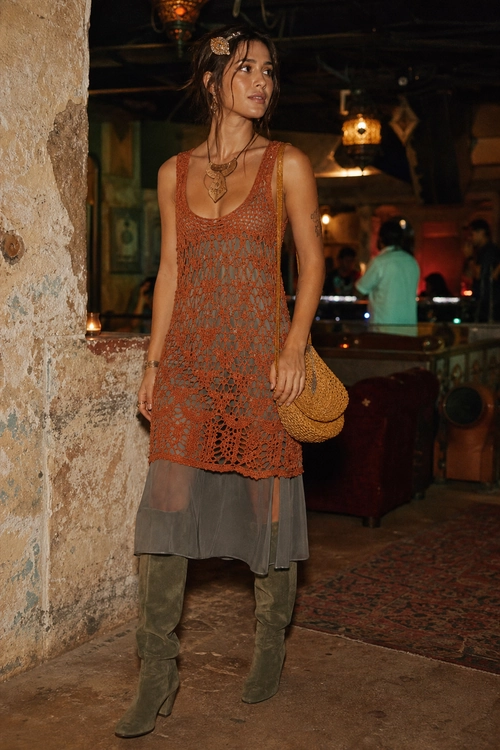 A layered outfit featuring a rust crochet sleeveless tunic, gray skirt, olive knee-high boots, and a woven shoulder bag.