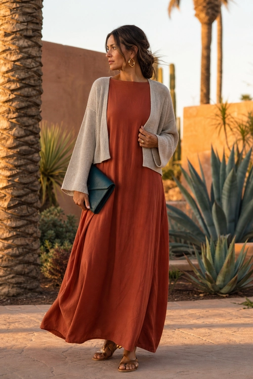 Rust Maxi Dress with Cropped Cardigan A woman wears a flowing rust maxi dress with a beige cropped cardigan, dark teal clutch, hoop earrings, and brown sandals.