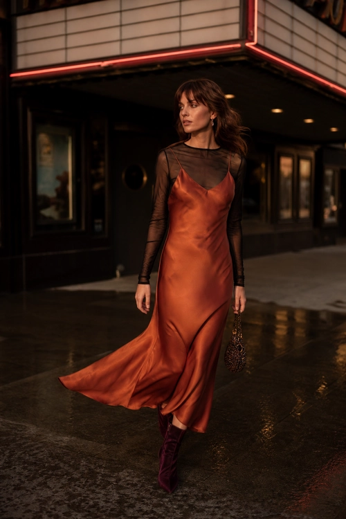Rust Satin Slip Dress Outfit A rust satin slip dress is layered over a sheer black long-sleeve top and styled with burgundy boots and a small beaded handbag.