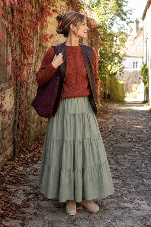 Rust Sweater and Tiered Skirt Outfit A layered outfit featuring a rust cable-knit sweater, dark brown vest, sage green tiered maxi skirt, burgundy tote, and beige shoes.