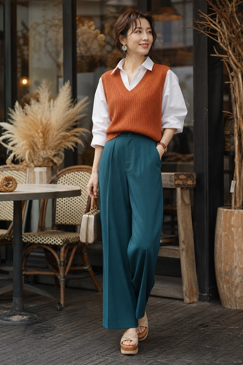 Rust Sweater Vest and Teal Trousers Outfit A woman wears a rust ribbed sweater vest over a white collared shirt with teal wide-leg trousers, platform sandals, and a small beige handbag.