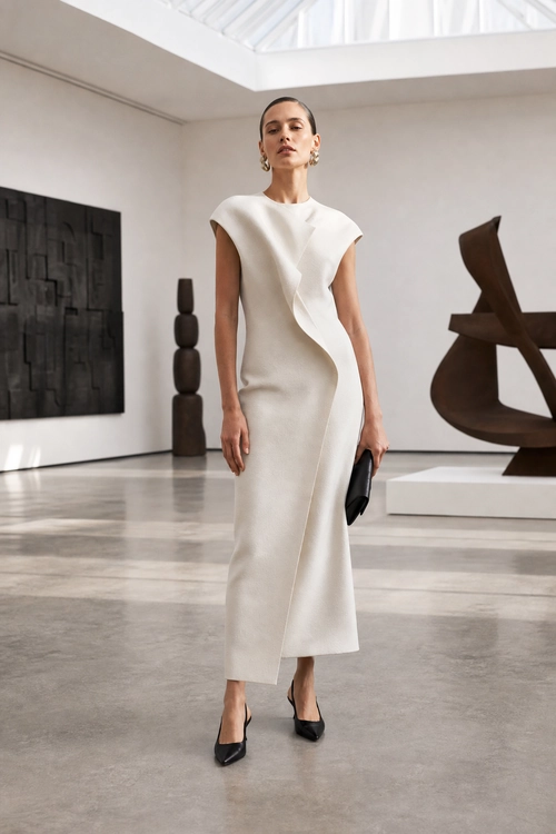 Sculptural Ivory Midi Dress A woman wears an ivory sleeveless midi dress with a sculptural asymmetric ruffle, black pointed heels, a black clutch, and gold earrings.