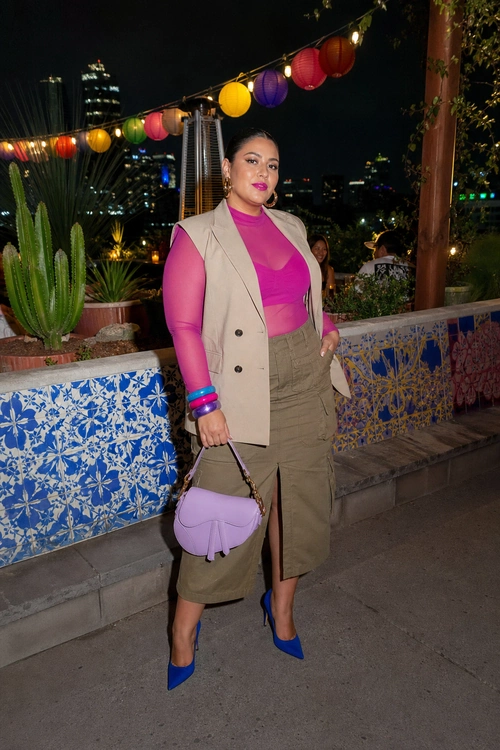 A woman wears a magenta sheer long-sleeve top with a beige sleeveless blazer, olive midi skirt, blue pointed heels, and a lavender handbag.