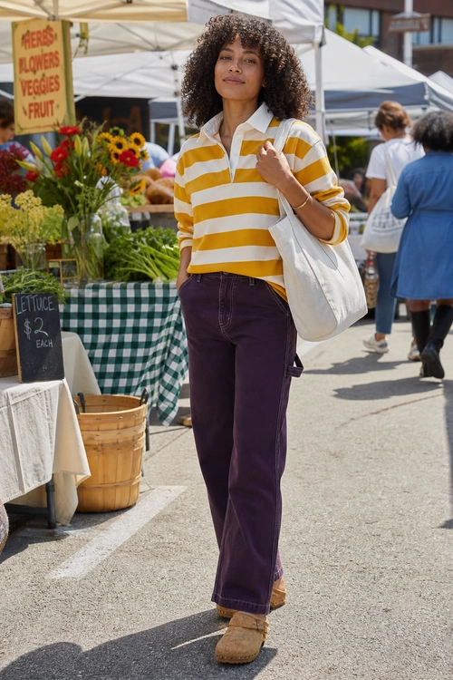 A woman wears a mustard-and-white striped collared shirt with purple pants, tan clogs, and a white tote bag at an outdoor market.