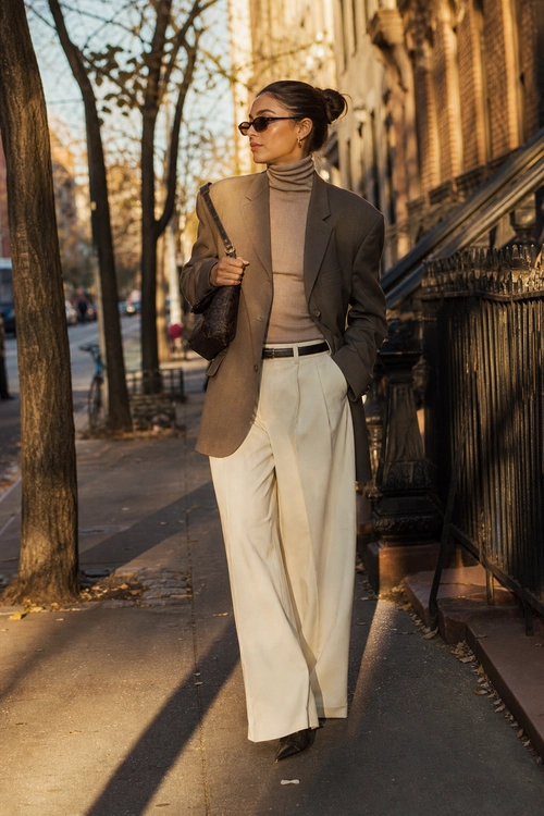 A woman wears a taupe blazer over a beige turtleneck with cream wide-leg trousers, a black belt, sunglasses, and a dark shoulder bag.
