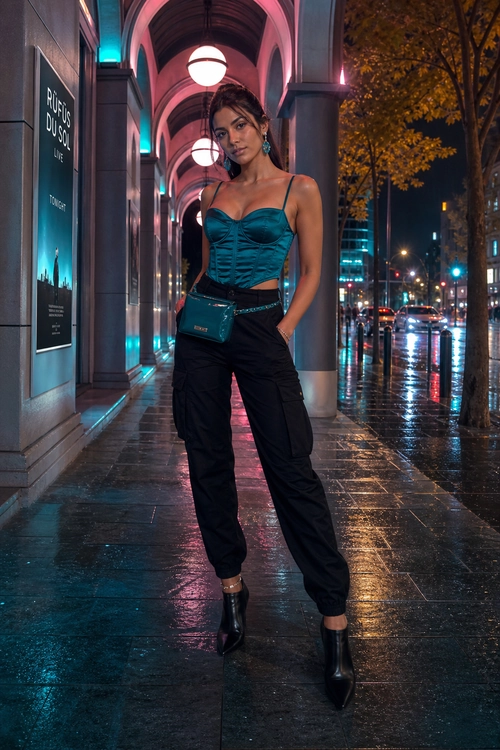 A woman wears a teal corset-style top with black cargo pants, ankle boots, and a small teal crossbody bag at night.
