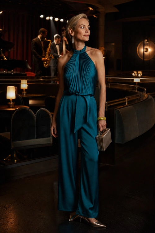 A woman wears a teal pleated halter jumpsuit with a tie waist, metallic pointed-toe pumps, drop earrings, a gold bracelet, and a box clutch.