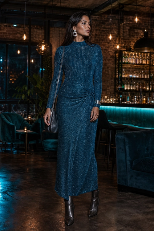 A woman wears a teal textured long-sleeve midi dress with a gathered waist, dark ankle boots, a shoulder bag, and drop earrings.