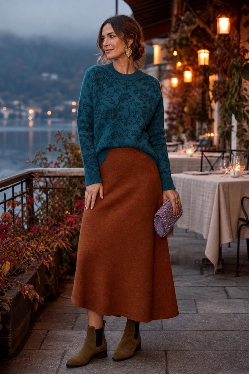 A woman wears a teal patterned sweater with a rust midi skirt, ankle boots, gold earrings, and a purple clutch.