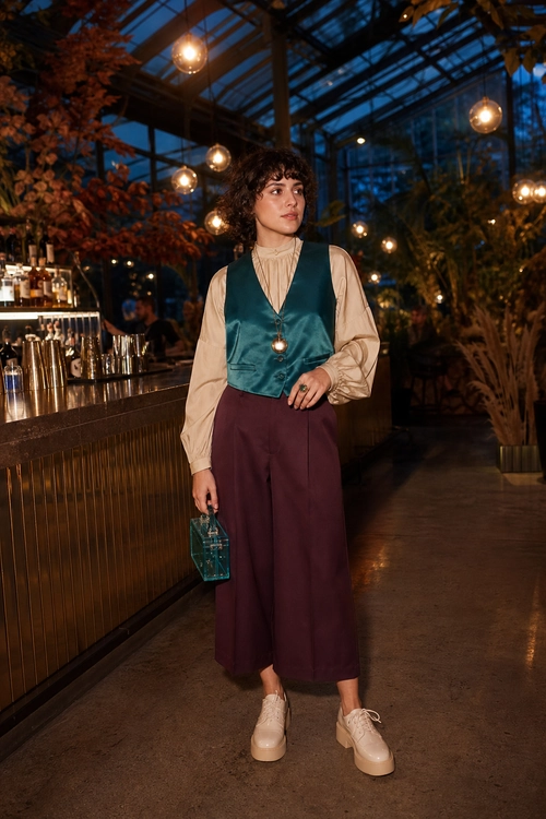 A fashion look featuring a teal satin vest over a beige blouse, plum wide-leg trousers, cream platform lace-up shoes, and a small teal handbag.