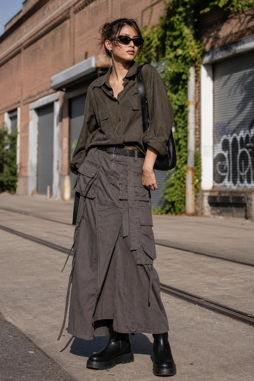 A street style outfit featuring a dark utility shirt, long cargo skirt, black sunglasses, shoulder bag, and chunky ankle boots.