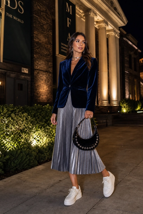 Velvet Blazer and Pleated Skirt Outfit Woman wearing a navy velvet blazer with a silver pleated midi skirt, white platform sneakers, and a black embellished handbag at night.