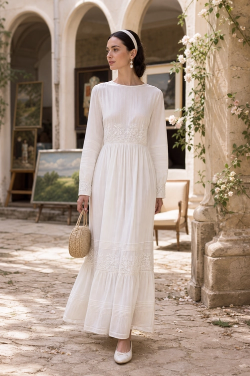 Woman wearing a white long-sleeved maxi dress with a black headband, drop earrings, woven handbag, and white flats.
