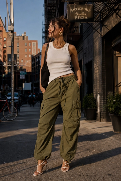 A woman wears a white sleeveless tank top with olive green cargo pants, strappy heeled sandals, gold jewelry, and a black shoulder bag on a city street.