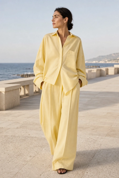Yellow Monochrome Coastal Outfit A woman wears a pale yellow button-up shirt with wide-leg matching trousers and flat sandals along a seaside promenade.