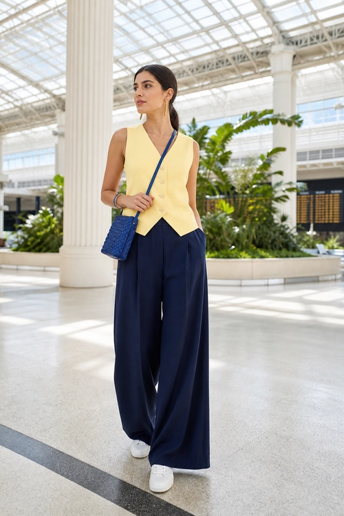 Yellow Vest and Navy Wide-Leg Trousers A woman wears a sleeveless yellow button-front vest with navy wide-leg trousers, white sneakers, and a blue crossbody bag.