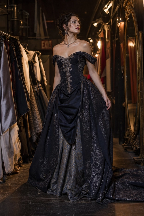 A woman wears a dramatic black off-shoulder ball gown with a corset-style bodice, full draped skirt, and long train.