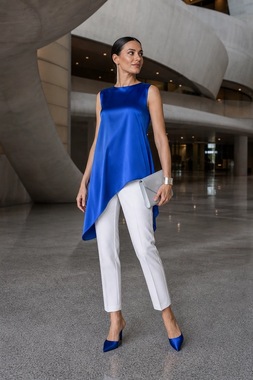 Woman wearing a royal blue sleeveless asymmetric tunic with white ankle-length pants, blue pointed-toe heels, a gray clutch, and a silver cuff bracelet.