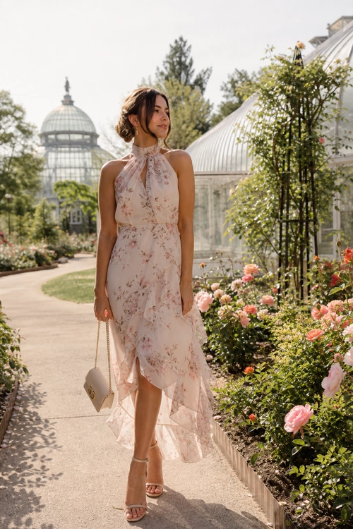 A woman wears a blush floral halter midi dress with nude strappy sandals and a small beige chain bag in a garden.
