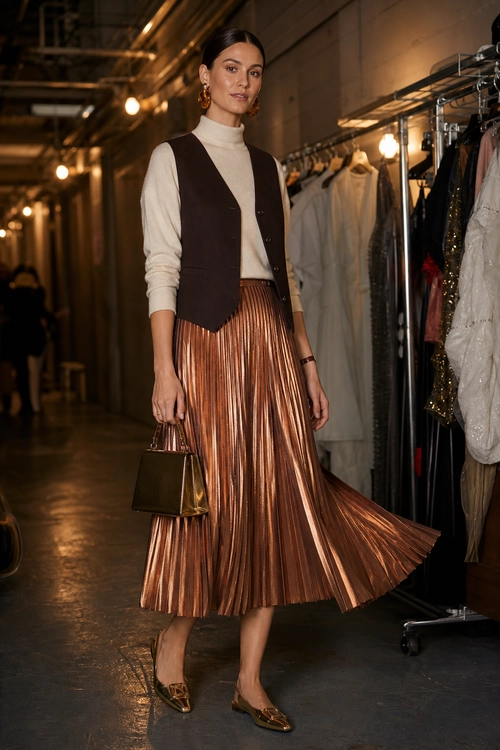A woman wears a cream turtleneck, dark brown buttoned vest, copper metallic pleated midi skirt, pointed flats, and carries a structured brown handbag.