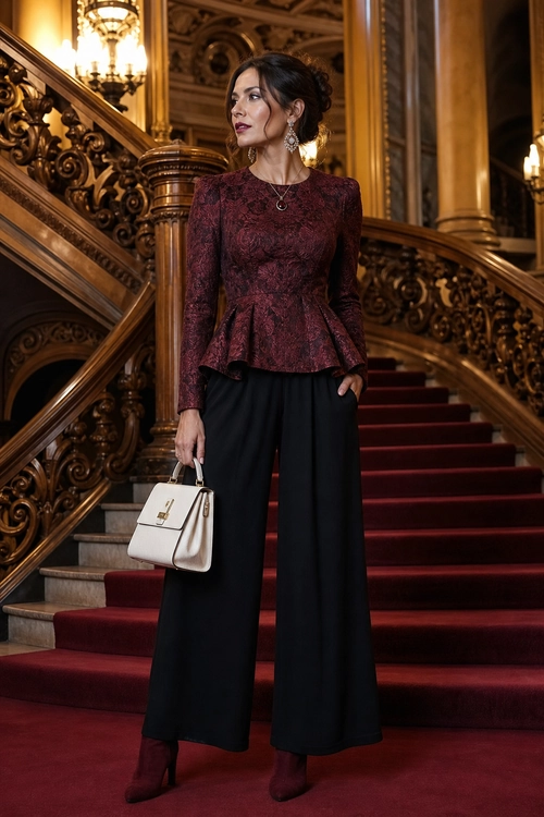 A woman wears a burgundy long-sleeve peplum top with black wide-leg trousers, burgundy ankle boots, and a cream structured handbag on an ornate staircase.