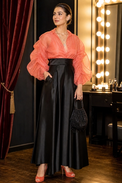 A woman wears a coral sheer blouse with voluminous sleeves, a high-waisted black leather skirt, coral open-toe heels, and a black beaded handbag.