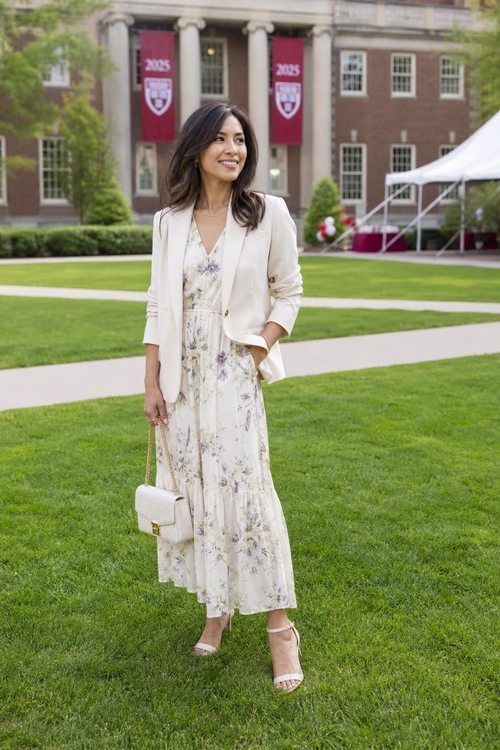 A woman wears a cream blazer over a light floral dress with heeled sandals and a small chain-strap handbag.
