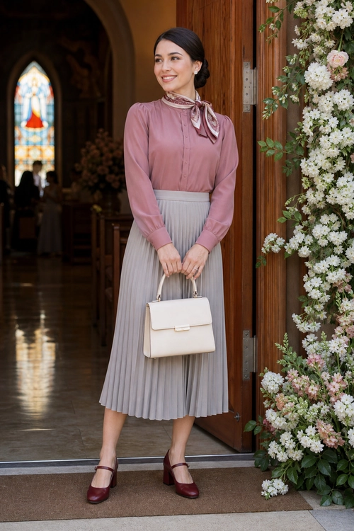 A woman wears a dusty rose long-sleeve blouse with a neck scarf, a gray pleated midi skirt, burgundy Mary Jane heels, and a cream handbag.