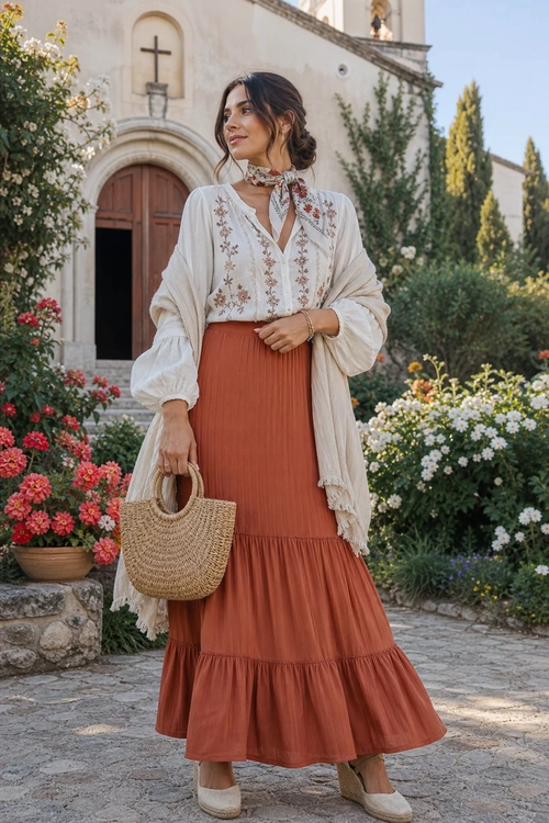 A woman wears an embroidered white blouse, rust tiered skirt, fringed cream shawl, woven tote, and espadrille shoes in a flower-filled courtyard.