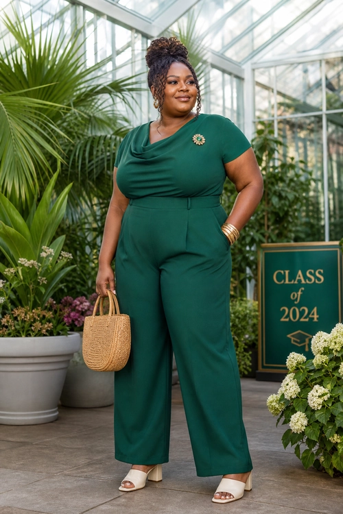 A woman wears an emerald green wide-leg jumpsuit with a brooch, woven handbag, gold bangles, and cream open-toe sandals.