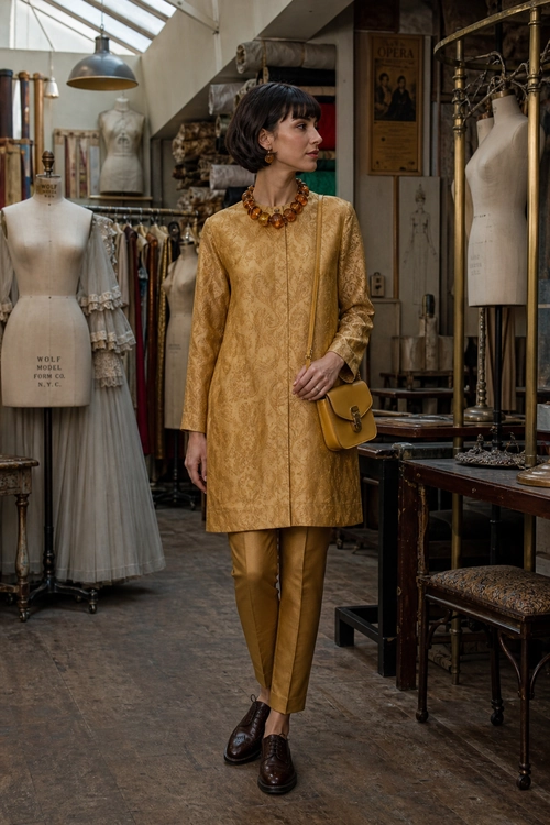 A woman wears a gold brocade tunic with matching trousers, brown loafers, a mustard crossbody bag, and an amber statement necklace.
