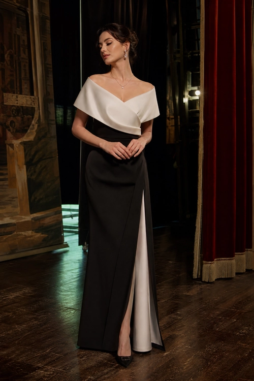 Ivory and Black Evening Gown A woman wears an ivory off-the-shoulder top with a black floor-length skirt featuring a high slit and light panel, styled with pointed heels and delicate jewelry.