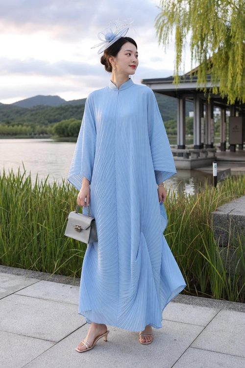 A woman wears a flowing light blue pleated maxi dress with a matching fascinator, structured handbag, and heeled sandals.