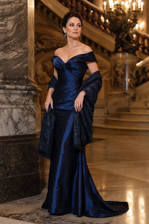 A woman wears a navy off-shoulder satin evening gown with a dark textured shawl and delicate jewelry.
