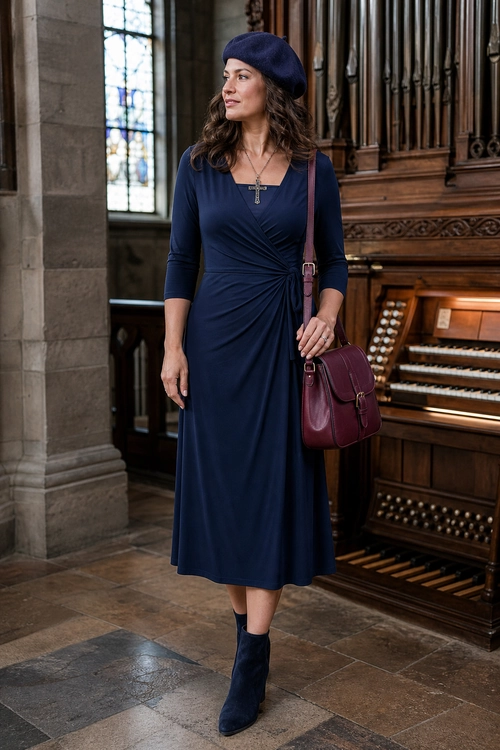 A woman wears a navy long-sleeve wrap dress with a matching beret, ankle boots, and a burgundy shoulder bag.