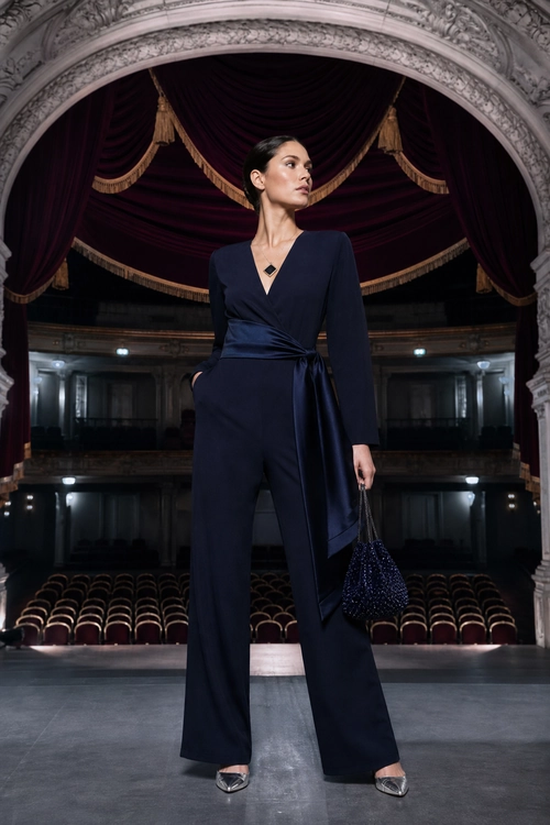 A woman wears a navy long-sleeve wrap jumpsuit with wide legs, silver pointed heels, a dark pendant necklace, and a beaded handbag.