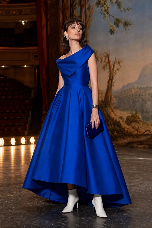 A woman wears a royal blue draped evening dress with a full asymmetric skirt, white pointed ankle boots, and a matching clutch.