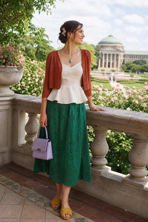 A woman wears a rust cropped cardigan over a cream top with a green eyelet midi skirt, woven sandals, and a lavender handbag on a garden terrace.