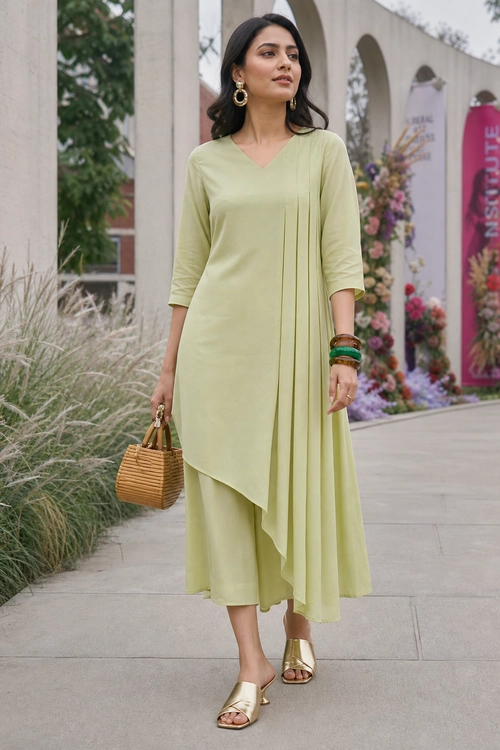 Sage Green Asymmetric Kurta Set Woman wearing a pale sage green asymmetric tunic with matching cropped wide-leg trousers, gold sandals, a woven handbag, hoop earrings, and stacked bangles.